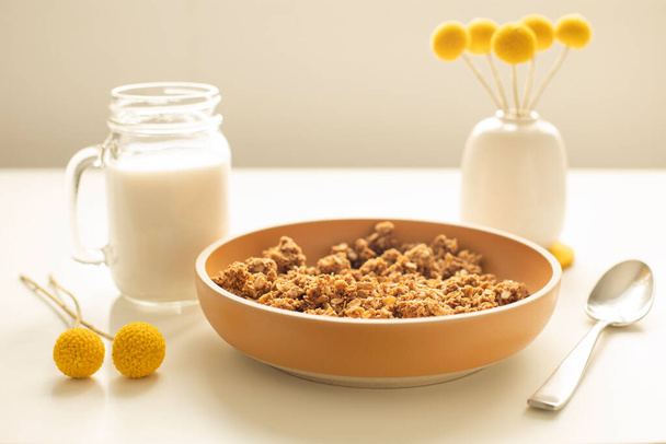 Side view of a yellow ceramic bowl with granola with a metal spoon, glass mug with almond milk, small vase with yellow flowers next to it, on white - Foto, imagen