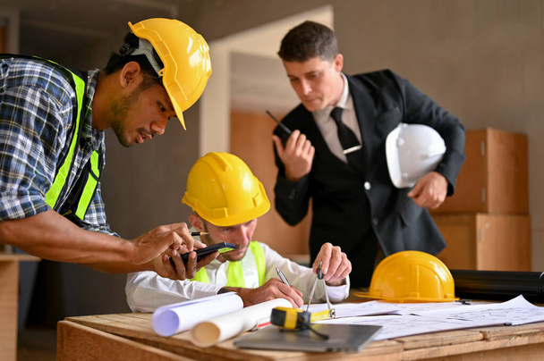 A professional Asian male construction inspector using scientific calculator while a caucasian male engineer using divider compass to measure scale on a blueprint. - Foto, Imagen