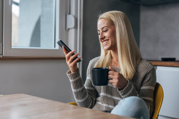 Beautiful woman looks at her phone and enjoys coffee in the kitchen - Zdjęcie, obraz