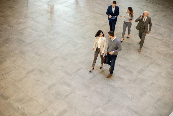 Group of young and senior business people are walking in an office hallway, captured in an aerial view. They are dressed in formal attire, walking with purpose and intent and discussing amongst themselves and interacting with colleagues - 写真・画像