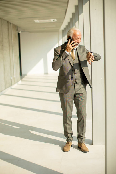 Un hombre de negocios de alto rango está en el pasillo de una oficina, centrado en su teléfono móvil. Está vestido con vestimenta formal, exudando confianza y profesionalismo. - Foto, imagen