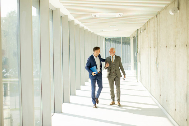 Young and a senior businessman walk down an office hallway, deep in conversation. They are both dressed professionally, reflecting their business acumen and status. They are navigating the complexities of the corporate world, finding solutions and ma - Foto, Imagem