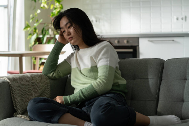 Aggódó ázsiai Y-generációs lány ül a kanapén, magányosnak érzi magát, monopóliumban szenved. Szomorú, depressziós, fiatal koreai nő otthon ül egyedül, félrenéz, és problémái vannak a pánikroham kezelésével. - Fotó, kép