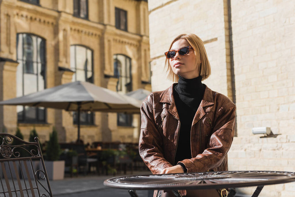 femme blonde en lunettes de soleil et veste en cuir élégant assis dans un café extérieur  - Photo, image