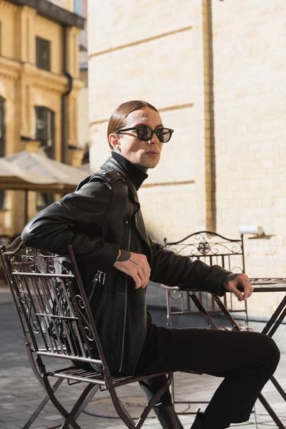 Татуированный молодой человек в стильных солнцезащитных очках и черной кожаной куртке сидит в кафе на открытом воздухе  - Фото, изображение