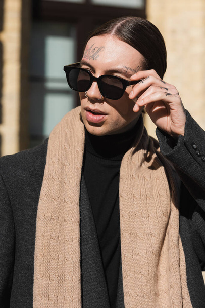 татуированный молодой человек в пальто и шарфе, регулирующий стильные солнечные очки - Фото, изображение