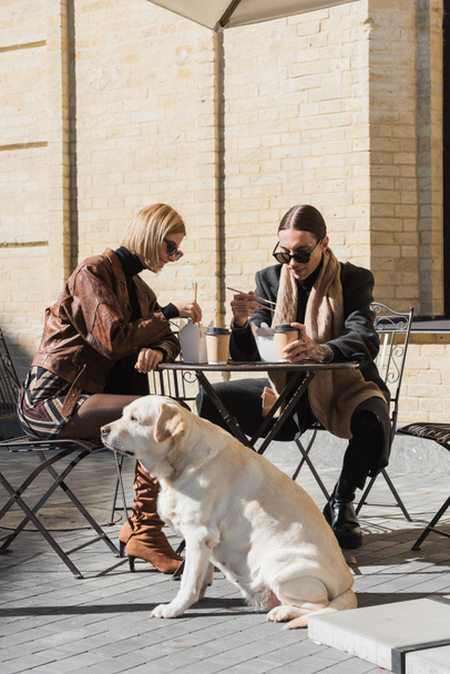 happy couple eating asian food near coffee to go while sitting with labrador in outdoor cafe  - Foto, imagen
