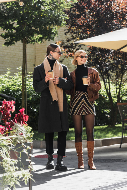 full length of stylish couple in sunglasses holding paper cups while standing near outdoor cafe  - Foto, Imagen