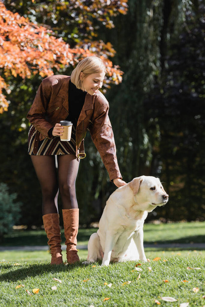 full length of happy blonde woman holding paper cup and cuddling labrador in park  - Φωτογραφία, εικόνα