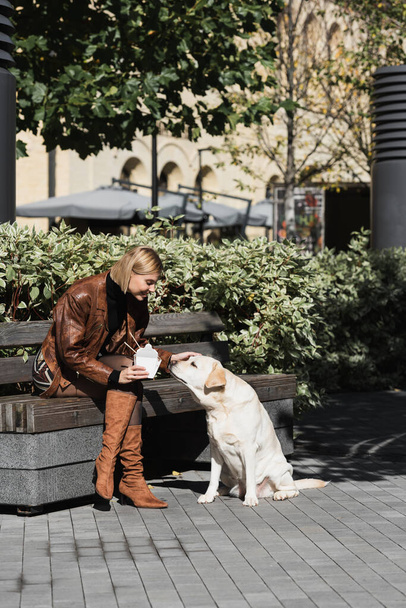 full length of smiling blonde woman in leather jacket holding takeaway box with asian food and cuddling dog  - Photo, Image