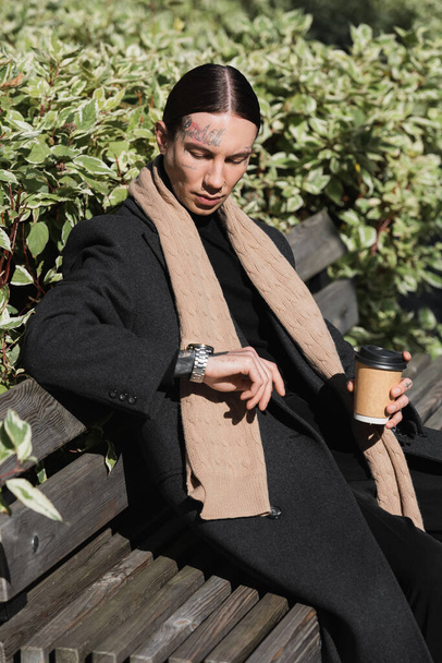 giovane uomo tatuato in cappotto e sciarpa che tiene bevanda da asporto mentre guarda l'orologio da polso e seduto sulla panchina  - Foto, immagini