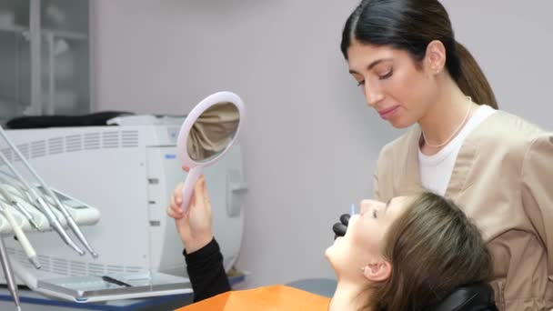 若い患者が歯の椅子に座っている。女性歯科医は歯科用鏡を持っている女性患者を扱います - 映像、動画