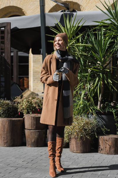 full length of happy blonde woman in beige coat and hat holding paper cup with coffee to go  - Foto, imagen