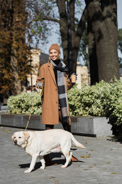 longitud completa de la mujer alegre en sombrero y abrigo sosteniendo taza de papel mientras pasea con el perro  - Foto, imagen