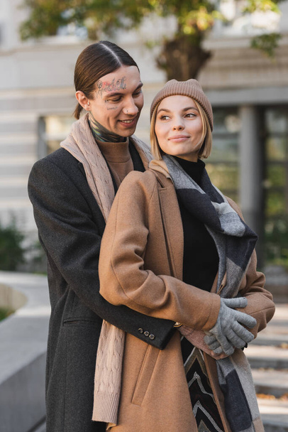 getatoeëerde man knuffelen blij blond vrouw in hoed terwijl staan buiten  - Foto, afbeelding