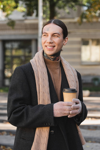 стильный мужчина в пальто и шарфе улыбается, держа бумажную чашку с кофе, чтобы пойти  - Фото, изображение
