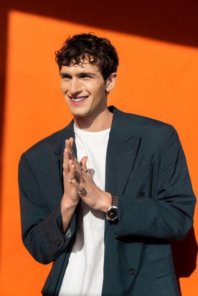 Cheerful and stylish man in jacket posing on orange background with sunlight  - Photo, Image