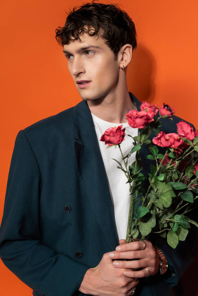 Stylish brunette man in blazer holding roses on orange background  - Фото, изображение