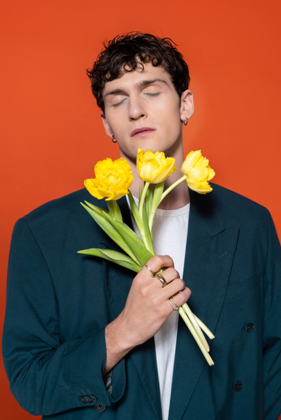 Brunette man in jacket closing eyes and holding yellow tulips isolated on red  - Foto, immagini