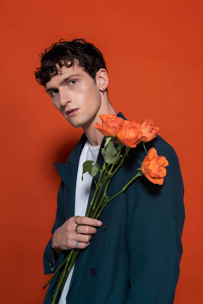 Trendy brunette man in blue suit holding orange flowers for 8 march on red background  - Foto, immagini