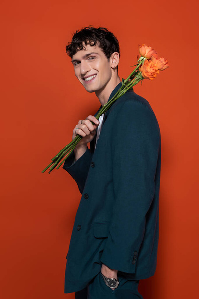 Smiling man in blue suit holding orange flowers for 8 march on red background  - Photo, Image