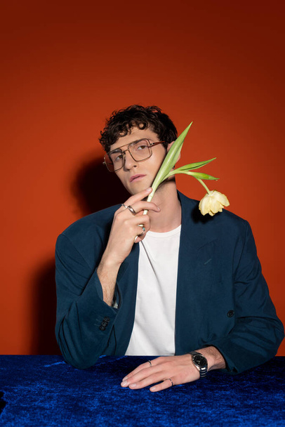 Young man in stylish outfit and eyeglasses holding tulip on red background - Foto, Imagem