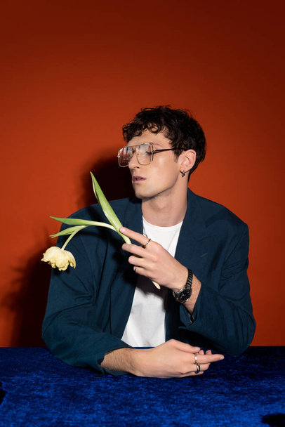 Fashionable curly model in eyeglasses holding tulip on red background - Fotó, kép
