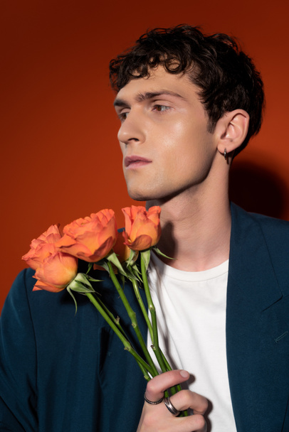 Portrait of stylish young man holding orange roses on red background - 写真・画像