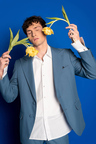 Trendy and curly young man holding tulips and closing eyes on blue background  - Foto, immagini