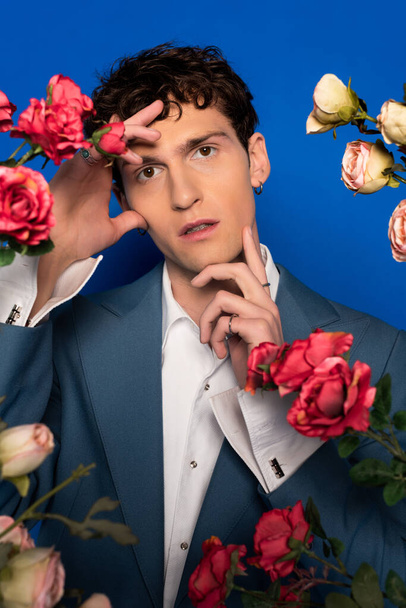 Portrait of stylish curly model in shirt and jacket touching face near roses on blue background  - Photo, Image