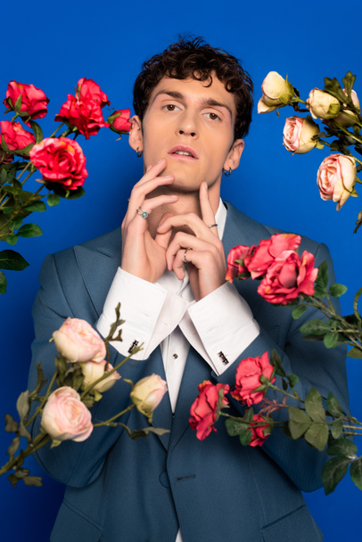 Curly man in stylish outfit touching chin near roses on blue background  - Foto, Imagem