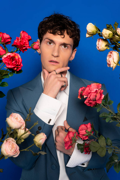 Curly man in stylish outfit touching chin near blooming flowers on blue background  - Valokuva, kuva