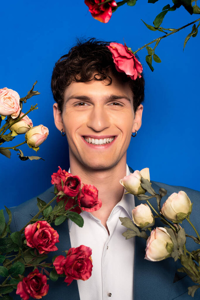 Портрет усміхненого чоловіка в елегантному одязі, який дивиться на камеру біля квітів на синьому фоні
  - Фото, зображення