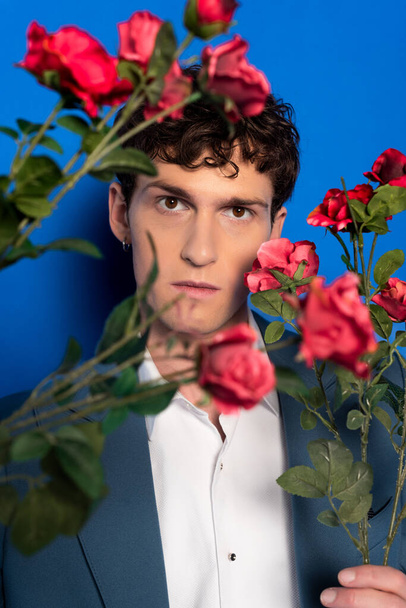 Portrait of brunette man in jacket and shirt holding flowers on blue background  - Photo, Image