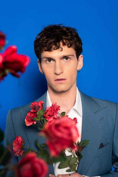 Curly man in jacket looking at camera near red flowers on blue background  - Photo, Image