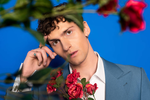krullend en stijlvol jongeman kijken naar camera in de buurt van rozen op blauwe achtergrond  - Foto, afbeelding