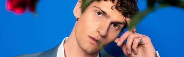 Trendy and curly man looking at camera near blurred flower on blue background, banner  - Photo, Image
