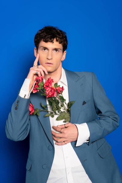 Modelo elegante na camisa e jaqueta segurando flores e posando no fundo azul  - Foto, Imagem