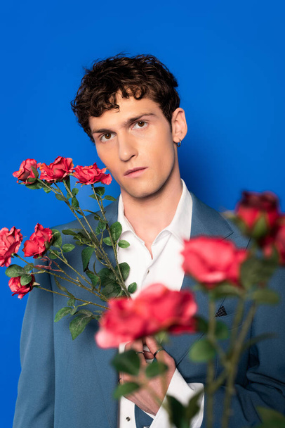 Portrait of curly young man in jacket posing with red flowers isolated on blue  - Фото, изображение