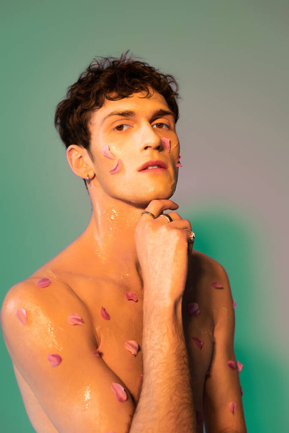 Hombre sin camisa con pétalos florales en la piel mirando a la cámara en un fondo colorido  - Foto, imagen