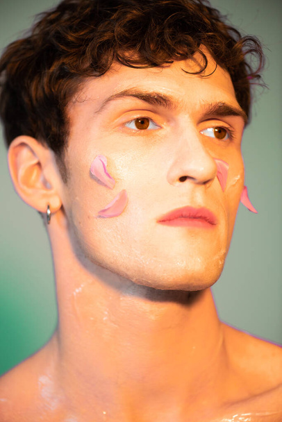 Yüzünde taç yaprakları olan genç bir adamın portresi renkli bir arkaplana bakıyor.  - Fotoğraf, Görsel