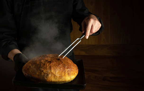 Baker bakt brood in de bakkerij. Vork in de hand van de chef en een bakplaat met warm brood. Plaats voor reclame op een donkere achtergrond - Foto, afbeelding