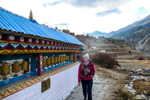 A woman walking along a wall full of prayer wheels on Annapurna Circuit Trek in Himalayas, Nepal. The woman enjoys the trek. There is a high Himalayan chain in the back. Spirituality and meditation - Foto, afbeelding
