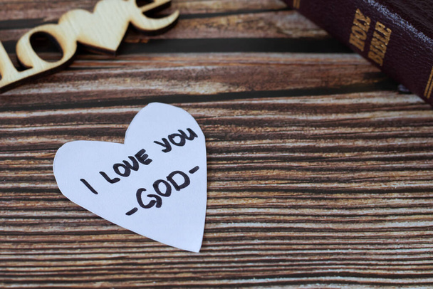 I love you-God, handwritten text on heart-shaped note and closed holy bible book on a wooden table. A closeup. Christian concept of love and hope in Jesus Christ. - Foto, immagini