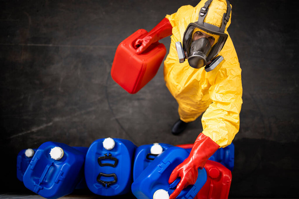 Bovenaanzicht van een beschermde werknemer in geel hazmatpak en gasmasker die in de chemische industrie werkt en plastic bussen met zuur neemt. - Foto, afbeelding