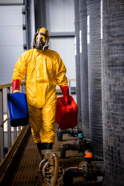 Chemicals worker carrying canisters and walking by large acid reservoir inside factory. - Foto, Imagen