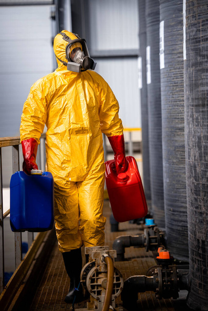 Experienced worker in yellow protection suit and gas mask working in chemicals factory and walking by large acid tanks. - Foto, Imagen