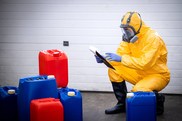 Trabajador de fábrica que usa traje de protección y máscara de gas comprobando inventario de ácido dentro del almacén. - Foto, imagen