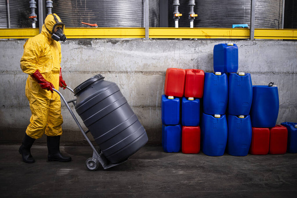 Professional worker in hazmat suit and gas mask carrying large barrel with acid in chemicals production factory. - Fotoğraf, Görsel