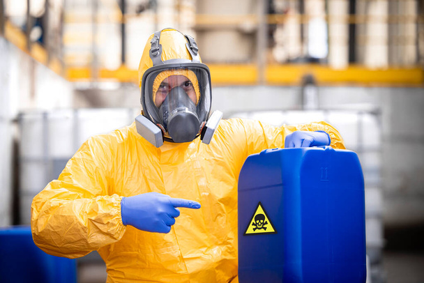 Fabrieksarbeider in beschermende apparatuur en gasmasker wijzend vinger naar de plastic blik met giftige chemicaliën. - Foto, afbeelding